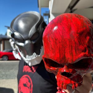 Może przedstawiać: Czarna i srebrna maska Predatora oraz czerwona czaszka. Czerwona czaszka ma błyszczące wykończenie i jest trzymana przed osobą noszącą czarny t-shirt z czerwoną grafiką. Maska Predatora jest w tle.