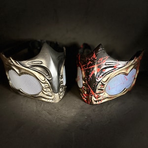 May include: Two gold-colored masks with intricate designs. One mask has a black base with red splatters, the other has a silver base. Both masks have a mesh covering the eye holes.