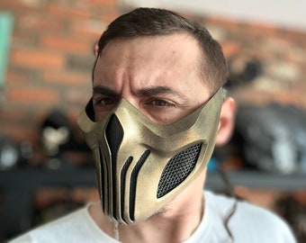 May include: A gold-colored mask with a skull-like design and a mesh covering the eyes. The mask is worn by a person with short brown hair.