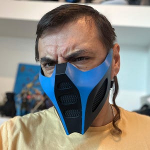 May include: A blue and black face mask with a mesh design over the mouth and nose. The mask is a cosplay item, likely inspired by a video game or comic book character.