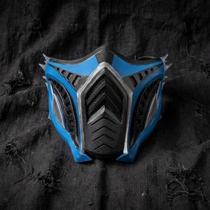 Puede incluir: Una máscara azul y negra con un diseño futurista. La máscara tiene rasgos afilados y angulares y una superficie texturizada. La combinación de colores incluye tonos de azul, negro y plata, creando un contraste visual llamativo. La máscara parece estar hecha de materiales duraderos.