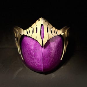 Può includere: Una maschera dorata ornata con un volto viola strutturato. La maschera presenta un design a forma di corona nella parte superiore e strutture a forma di ali sui lati. Il volto viola ha un aspetto screpolato e strutturato, su sfondo scuro.