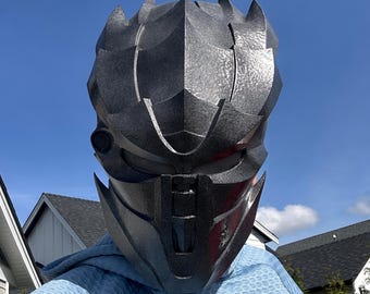 May include: A dark gray, textured, full-face helmet with a sharp, angular, multi-layered design. The helmet features a prominent central ridge, recessed eye slits with mesh detailing, and a vertical nose guard, creating a rigid, armored appearance.