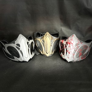 Könnte beinhalten: Drei schwarze, weiße und goldene Gesichtsmasken mit komplizierten Designs. Die Masken haben eine strukturierte, fast lederartige Oberfläche.