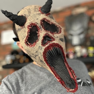 May include: A burlap mask with a skull design, red stitching, and black horns. The mouth is open and has a black and red jagged edge.