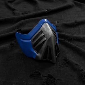 May include: A blue and gray mask with a futuristic design. The mask has a blue frame and a gray center with a mesh-like pattern. The mask is likely for cosplay or costume use.