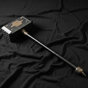 May include: A large, dark gray and bronze hammer with a long, black handle. The hammerhead features a decorative bronze emblem and silver accents. The handle has a pointed end. The hammer is set against a dark, textured background.