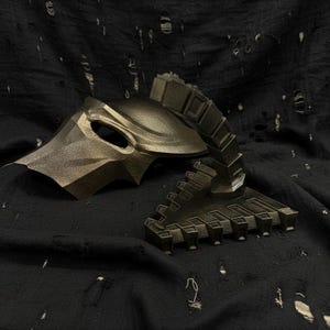 Könnte beinhalten: Eine metallisch goldfarbene Maske und ein segmentiertes, gegliedertes Teil, möglicherweise ein Helm- oder Kostümzubehör, werden vor einem dunklen, strukturierten Hintergrund gezeigt. Die Maske hat eine Augenöffnung und kantige Merkmale. Das segmentierte Teil ist aufrecht.