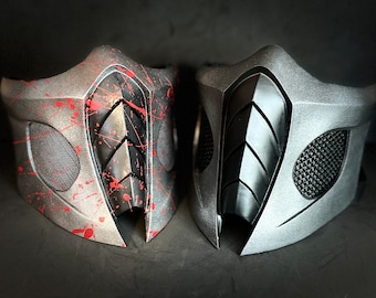 Puede incluir: Dos máscaras faciales, una con un diseño negro y plateado y la otra con un diseño rojo y plateado. Ambas máscaras tienen un patrón geométrico y están hechas de un material duro.