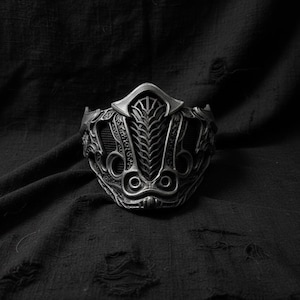 May include: A detailed, metallic mask with an intricate design. The mask features a central, vertical pattern and symmetrical, circular openings. The overall aesthetic is dark and gothic, with a focus on texture and detail.
