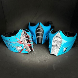 Könnte beinhalten: Drei blaue und schwarze Gesichtsmasken mit unterschiedlichen Designs. Eine Maske hat ein rotes Spritzmuster, eine hat ein schwarz-weißes geometrisches Muster und eine hat ein schwarz-weißes Glitzermuster.