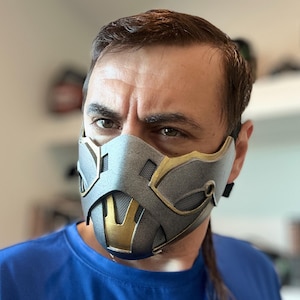 Könnte beinhalten: Eine grau-goldene Gesichtsmaske mit einem Netzabschnitt über dem Mund. Die Maske hat ein metallisches Aussehen und wird von einer Person getragen, die ein blaues Hemd trägt. Die Maske hat ein detailliertes, eckiges Design.