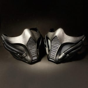 May include: Two metallic gray face masks with a futuristic design. Each mask features a contoured shape with mesh detailing and ribbed accents. The masks are displayed against a dark background, highlighting their intricate design.
