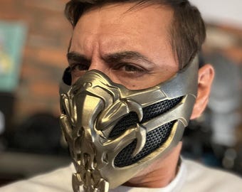 May include: A gold and silver mask with detailed, sculpted features. The mask has a mesh section for the mouth and nose, and a complex, stylized design. The mask is worn by a person.