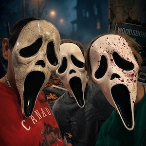 May include: Three Scream masks, a popular Halloween costume, are displayed. The masks are white with black eye and mouth openings. One mask has a cracked, aged appearance, while another is splattered with red paint. The person on the left is wearing a red shirt with the word "CANADA".