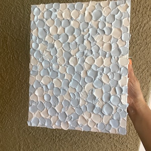 May include: Abstract artwork featuring a textured pattern of light blue and cream-coloured paint strokes. The square canvas is held by a hand, showcasing the tactile surface.