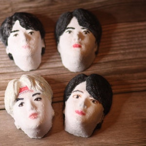 May include: Four white, sculpted face busts with painted features, including black hair, eyes, and red lips. One bust has silver hair and a red headband. The busts are arranged on a wooden surface.