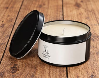 May include: A black metal candle tin with a matching lid placed to the side. The candle contains off-white wax with a single centered wick. A white label on the front features a black line drawing of hands holding a candle and the text HANDMADEE SOAP & CANDLE CO.