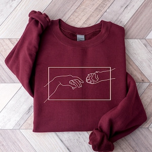 May include: A maroon sweatshirt with a white outline of a human hand and a dog paw reaching towards each other, reminiscent of the "Creation of Adam" painting.