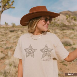 May include: An ivory short-sleeved t-shirt featuring two dark brown line-art sheriff star badges printed on the front. The right star includes the text "Amber's Bachelorette Nashville, 2026" written in a cursive script below the badge design.