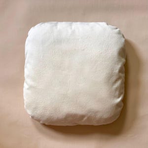 May include: A square, off-white pillow with a soft, textured surface. The pillow is a solid color and appears to be made of a plush material. The pillow is a neutral color and would complement a variety of home decor styles.