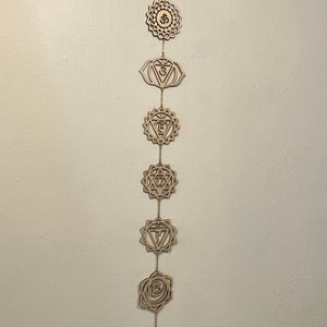 May include: A wooden chakra wall hanging with seven symbols, each connected by a chain. The top piece has an Om symbol. The symbols are in a natural wood color and are hanging against a white wall.