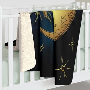 May include: A black and gold fleece throw blanket featuring a crescent moon and stars design. The moon is outlined in gold and has a textured surface. The stars are also gold and have a sparkly effect.