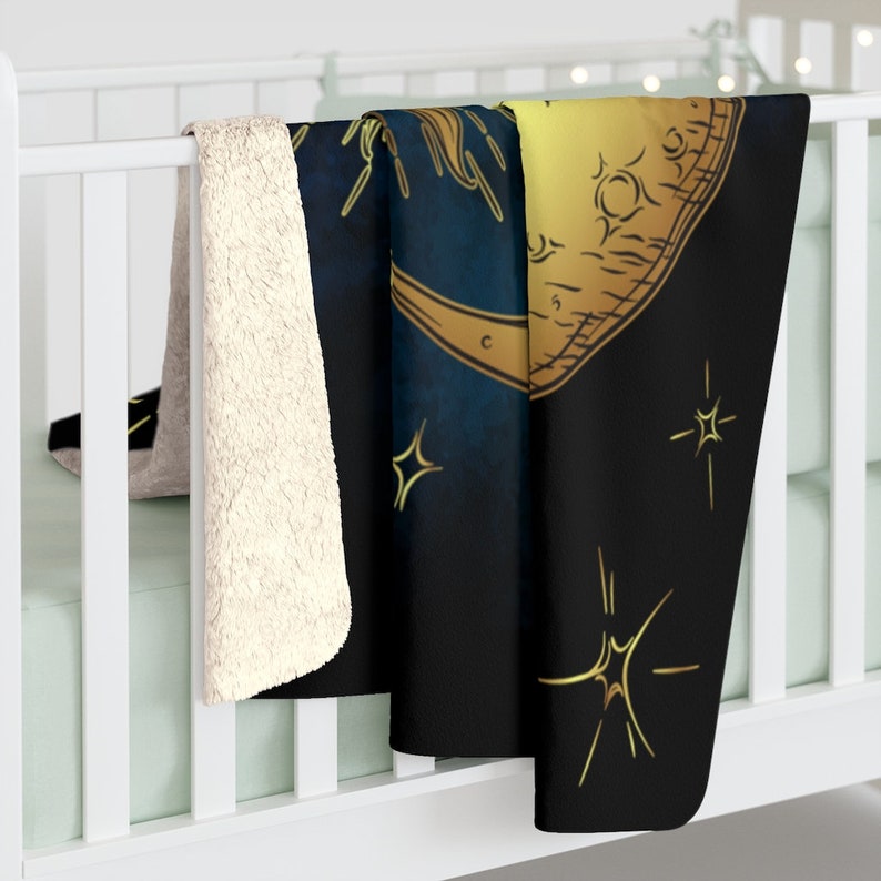May include: A black and gold fleece throw blanket featuring a crescent moon and stars design. The moon is outlined in gold and has a textured surface. The stars are also gold and have a sparkly effect.
