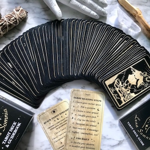 Op de afbeelding: Een zwart tarotdeck met gouden accenten en een handleiding. De kaarten zijn uitwaaierend gelegd om de achterkant te laten zien, die een halve maan en sterren heeft. De handleiding heeft een zwarte omslag met gouden letters die "Luna Somnia Tarot Deck & Guidebook" lezen.