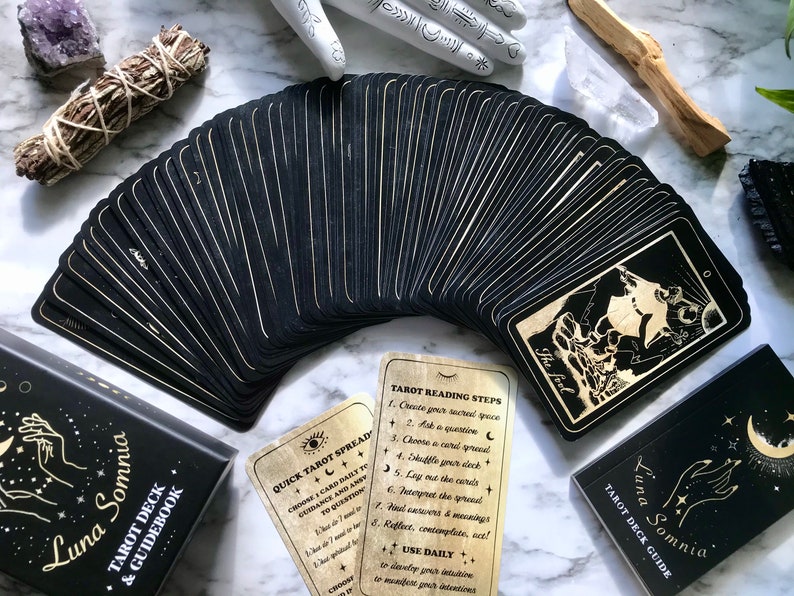 Op de afbeelding: Een zwart tarotdeck met gouden accenten en een handleiding. De kaarten zijn uitwaaierend gelegd om de achterkant te laten zien, die een halve maan en sterren heeft. De handleiding heeft een zwarte omslag met gouden letters die "Luna Somnia Tarot Deck & Guidebook" lezen.