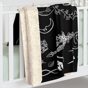 Op de afbeelding: Zwarte en witte fleece deken met een patroon van verschillende witte illustraties op een zwarte achtergrond. De illustraties omvatten een halve maan, een slang, een hand die een mes vasthoudt, een blad, een bot en een boeket bloemen.