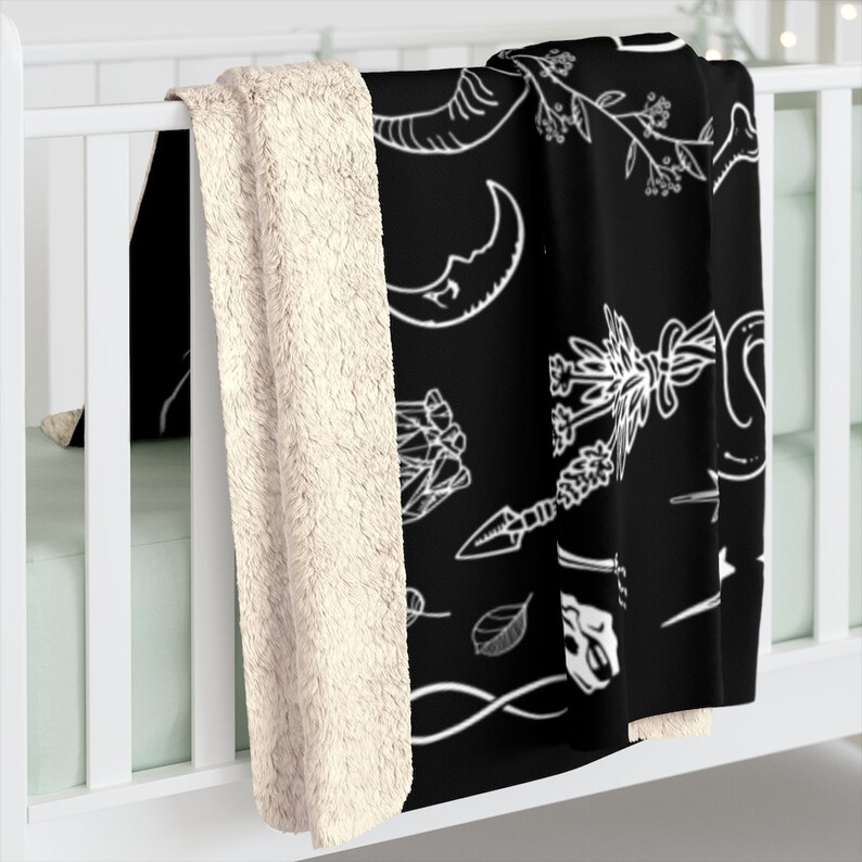 Op de afbeelding: Zwarte en witte fleece deken met een patroon van verschillende witte illustraties op een zwarte achtergrond. De illustraties omvatten een halve maan, een slang, een hand die een mes vasthoudt, een blad, een bot en een boeket bloemen.