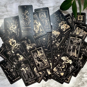 Op de afbeelding: Een set tarotkaarten met zwarte achtergronden en gouden illustraties. De kaarten liggen verspreid op een marmeren oppervlak.