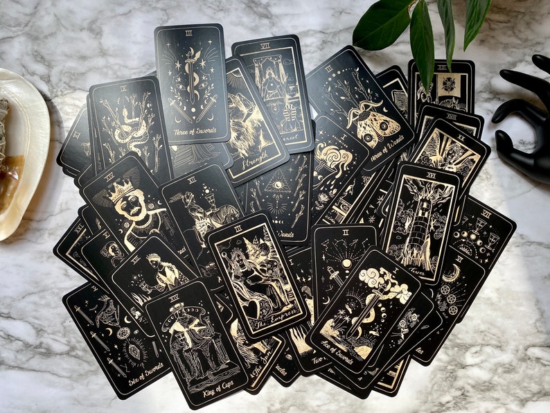 Op de afbeelding: Een set tarotkaarten met zwarte achtergronden en gouden illustraties. De kaarten liggen verspreid op een marmeren oppervlak.