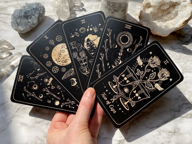 Op de afbeelding: Een set zwarte tarotkaarten met gouden accenten. De kaarten bevatten verschillende illustraties, waaronder een maan, sterren en een vrouw die een beker vasthoudt. De kaart op de voorgrond is de Twee van Bekers, die twee gouden bekers toont die met een roos zijn verweven.