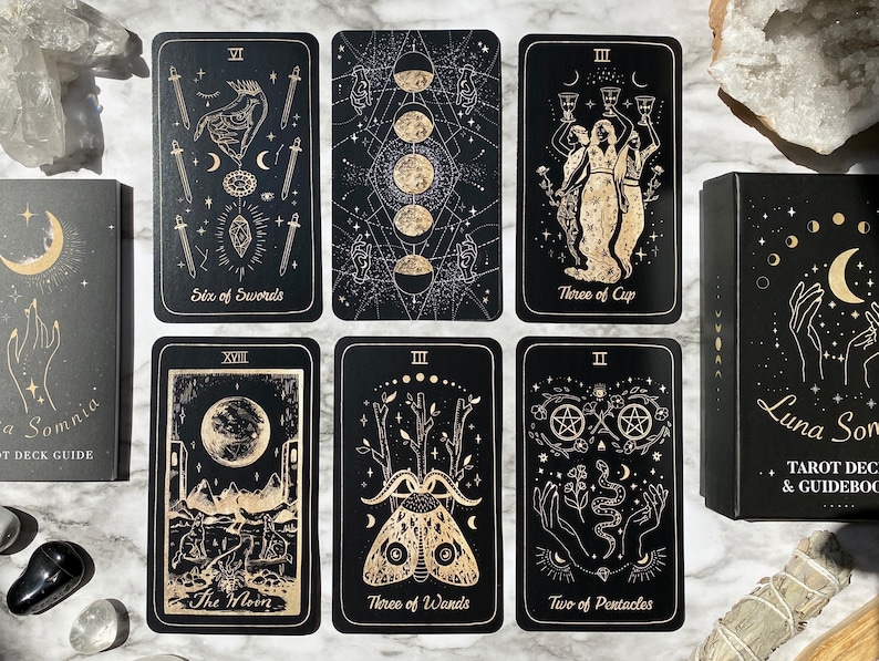 Op de afbeelding: Een zwarte tarotdeckdoos met gouden folielettering die "Luna Somnia Tarot Deck & Guidebook" luidt. De doos is open en onthult een set tarotkaarten met zwarte achtergronden en gouden folieillustraties. De kaarten zijn gerangschikt in twee rijen van drie, met de namen van de kaarten zichtbaar: Six of Swords, Three of Cups, The Moon, Three of Wands, Two of Pentacles.