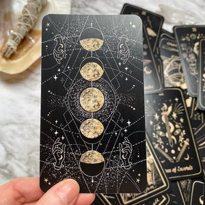 Op de afbeelding: Een zwarte tarotkaart met gouden foliedruk die de maanfasen voorstelt: sikkelmaan, halve maan, volle maan en wassende maan. De kaart is omringd door een geometrisch patroon van sterren en handen.