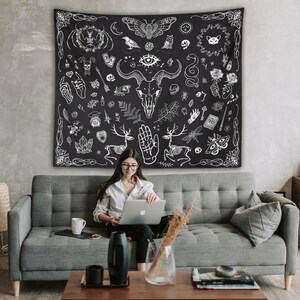 May include: A black and white tapestry with a variety of occult and mystical symbols, including a skull, a hand, a deer, a moth, and a snake. The tapestry is hanging on a wall behind a person sitting on a gray couch.