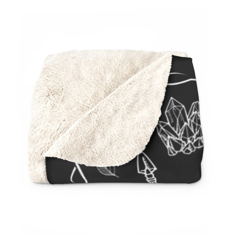 Op de afbeelding: Een zwart-witte deken met een ontwerp van kristallen, pijlen en bladeren. De deken heeft een zachte, pluche witte fleece aan de ene kant en een zwarte bedrukte kant.