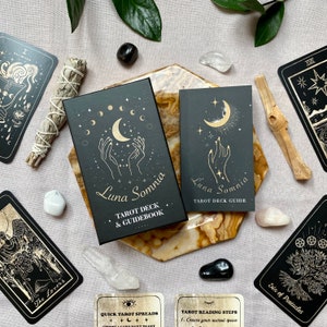 Op de afbeelding: Zwarte en gouden tarotdeckdoos met de titel "Luna Somnia Tarot Deck & Guidebook". De doos is omringd door kristallen, salie en andere tarotkaarten.