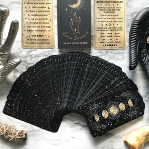 Op de afbeelding: Een zwart tarotdeck met gouden accenten en een hemels thema. De kaarten hebben een sterrenbeeldontwerp met manen en sterren. De handleiding bevat instructies voor het gebruik van het deck en een snelle tarotspread.