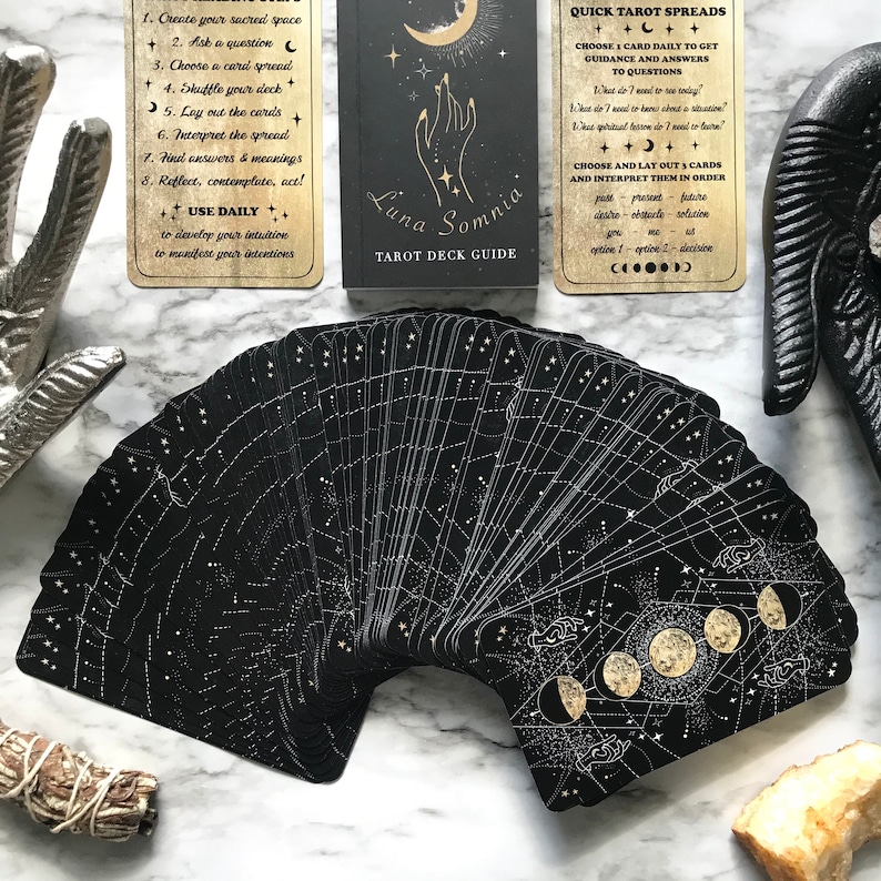 Op de afbeelding: Een zwart tarotdeck met gouden accenten en een hemels thema. De kaarten hebben een sterrenbeeldontwerp met manen en sterren. De handleiding bevat instructies voor het gebruik van het deck en een snelle tarotspread.