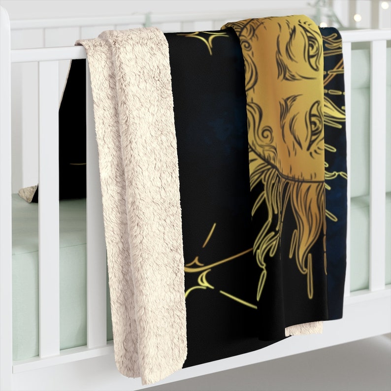 May include: A black and gold fleece throw blanket with a detailed illustration of a sun with a face and rays. The blanket is folded and draped over a white cot.