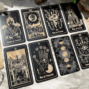 Op de afbeelding: Een tarotkaartendeck met zwarte achterkanten en gouden accenten. De kaarten bevatten illustraties van de Koningin van Zwaarden, de Maan, Negen van Stokken, Koning van Zwaarden, de Geliefden, Vijf van Pentakels en Zeven van Bekers.
