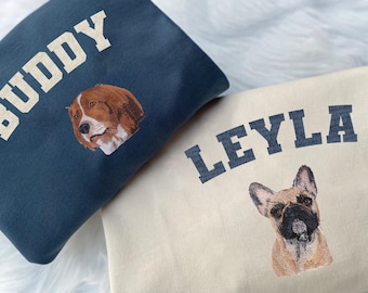 May include: Two sweatshirts, one navy blue and one cream, each embroidered with a dog portrait and a name. The navy sweatshirt says "BUDDY" in white block letters above a brown and white dog. The cream sweatshirt says "LEYLA" in navy block letters above a brown and black dog.