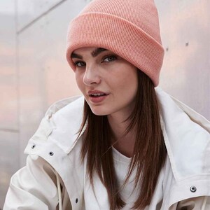 May include: A light peach-colored ribbed knit beanie hat. The hat is worn on the head and has a folded brim. The person is wearing a white jacket and a white shirt.