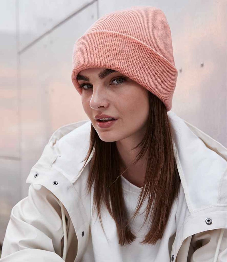 May include: A light peach-colored ribbed knit beanie hat. The hat is worn on the head and has a folded brim. The person is wearing a white jacket and a white shirt.
