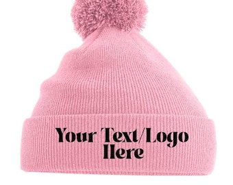 May include: A light pink beanie with a matching pom-pom on top. The folded brim of the hat displays the text "Your Text/Logo Here" in black. The hat is made of a ribbed knit material.