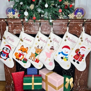 Può includere: Calze di Natale bianche con disegni ricamati di un pinguino, una volpe, una renna, una ballerina, un pupazzo di neve e Babbo Natale. Ogni calza ha un nome ricamato in rosso. I regali sono sotto.