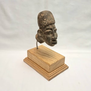 Puede incluir: Una escultura de cerámica marrón oscuro de una cabeza humana sobre un soporte de madera. La escultura tiene una cara detallada con la boca cerrada y una superficie texturizada.